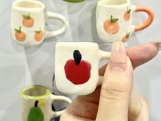 a hand holding a miniature coffee mug in front of several small cups with fruit painted on them