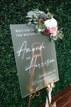 a sign with flowers on it sitting in front of a green wall that says, welcome to the wedding of amanda and harrison