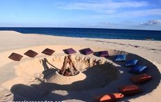 a fire pit made out of sand on the beach with colorful pillows around it and water in the background