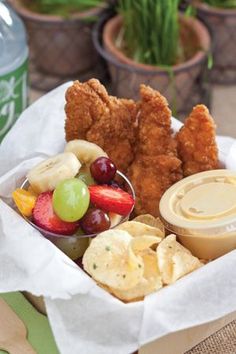 a basket filled with chicken, fruit and crackers next to a can of soda