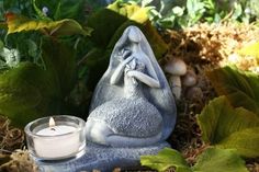 a candle is sitting on the ground next to some plants and rocks, with a statue of a person holding a baby