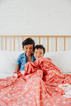 two children are laying on a bed under a red blanket and smiling at the camera