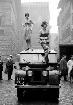 two women standing on top of an old truck in the middle of a crowded street