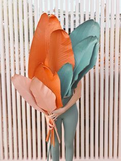 a woman holding a large bunch of orange and green leaves on her back while standing in front of a white fence