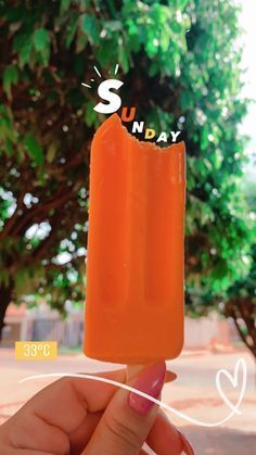 someone is holding an orange popsicle with the word sunday on it in front of some trees