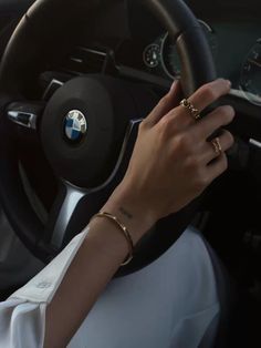 a woman driving a car with her hands on the steering wheel and wearing gold rings