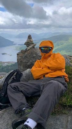 a man in an orange jacket sitting on top of a rock next to a backpack