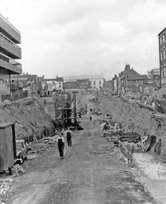 an old black and white photo of people walking down a dirt road with buildings in the background