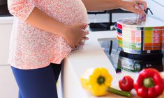 a pregnant woman standing in front of a counter with vegetables on it and measuring tape around her waist