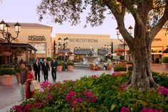 people are walking around in front of a shopping center with flowers on the ground and trees