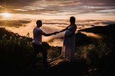 a man and woman holding hands on top of a mountain with clouds in the background