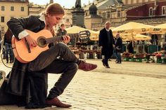 a woman sitting on the ground playing an acoustic guitar in front of people walking by