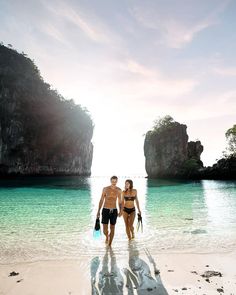 two people are walking on the beach in front of some rocks and water, while one person is holding a snorkel