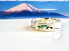 a silver ring with a green lizard on it's side and a mountain in the background