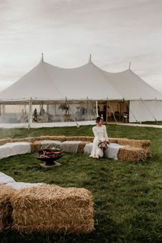 a woman sitting on hay bales in front of a large white tent and fire pit