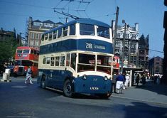 a double decker bus driving down a street next to tall buildings and people standing on the sidewalk