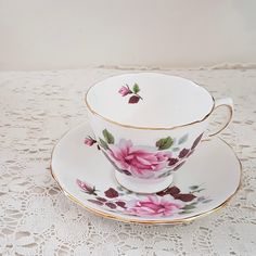 a tea cup and saucer with pink flowers on it sitting on a lace tablecloth