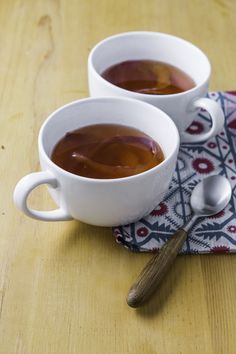 two white cups filled with brown liquid on top of a wooden table next to spoons