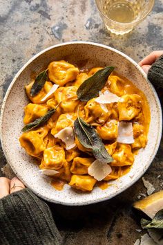 two hands holding a bowl of pasta with sage and cheese on the side next to a glass of wine