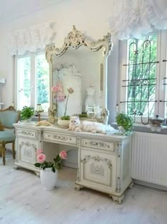 a white desk with a mirror and flowers on it in front of two large windows