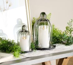 two white candles sitting on top of a table next to a mirror and greenery