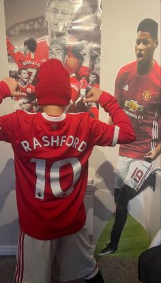 a young boy wearing a red hat and soccer jersey standing in front of a poster