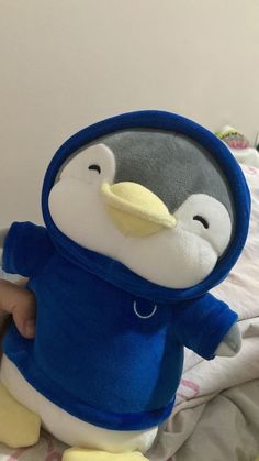 a stuffed penguin with a blue shirt and yellow feet is sitting on a white bed