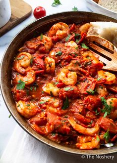 a skillet filled with shrimp and tomato sauce next to bread on a white table