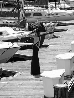 a woman in a long black dress standing on a dock next to several small boats