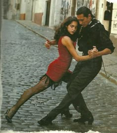 a man and woman dancing in the rain on a cobblestone street with buildings behind them