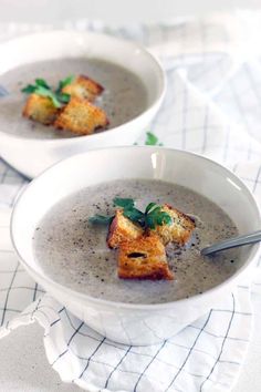 two white bowls filled with soup and croutons on top of a tablecloth