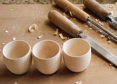 three white cups sitting on top of a wooden table next to some knives and wood shavings