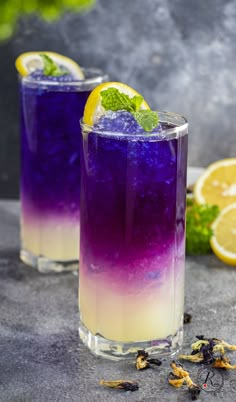 two glasses filled with purple and yellow liquid next to lemon slices on a gray surface