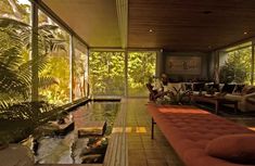 a living room filled with lots of furniture and plants next to a swimming pool in front of large windows