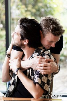 two men hug each other while sitting at a table in front of a large window