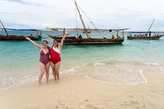 two women in bathing suits standing on the beach with boats in the water behind them