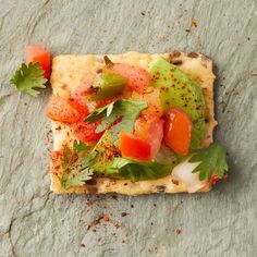 a square piece of food with tomatoes and herbs on the top, sitting on a stone surface