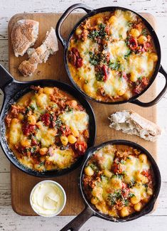 three cast iron skillets filled with food on top of a cutting board next to bread