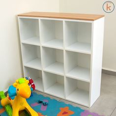 a toy giraffe sitting on the floor next to a white bookcase with lots of shelves