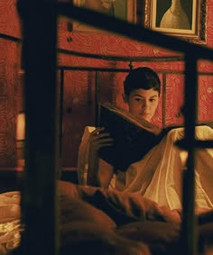 a young man reading a book in his bed with red wallpaper and paintings on the walls