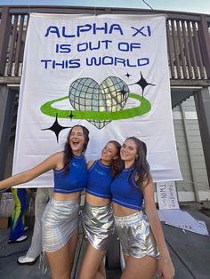 three girls in silver shorts and blue shirts posing for the camera with a banner behind them