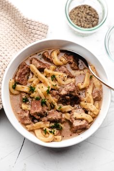 a bowl filled with beef and noodles on top of a white table next to a spoon