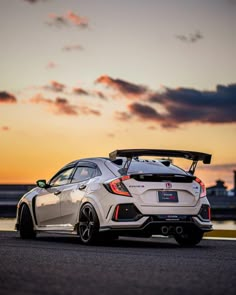 the rear end of a white sports car parked in front of an ocean at sunset