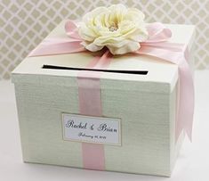 a white box with a pink ribbon and a flower on top that is attached to the lid