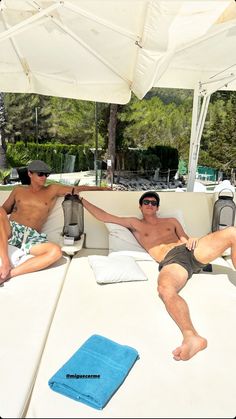 two men laying on top of a white couch next to each other under an umbrella