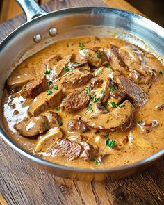 a pan filled with meat and mushrooms on top of a wooden table next to a spoon