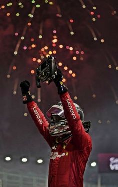 a man in a racing suit holding up his helmet with fireworks in the sky behind him