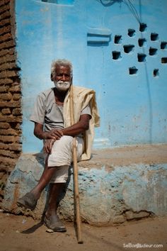 an old man sitting in front of a blue building with a wooden stick leaning on it