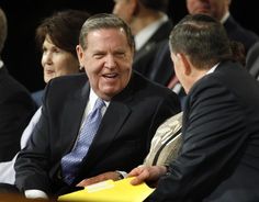 two men and one woman sitting at a table with other people in the background talking