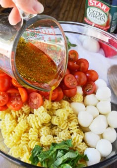 someone pouring dressing into a bowl filled with pasta and tomatoes, mozzarella shells, basil leaves, and cherry tomatoes